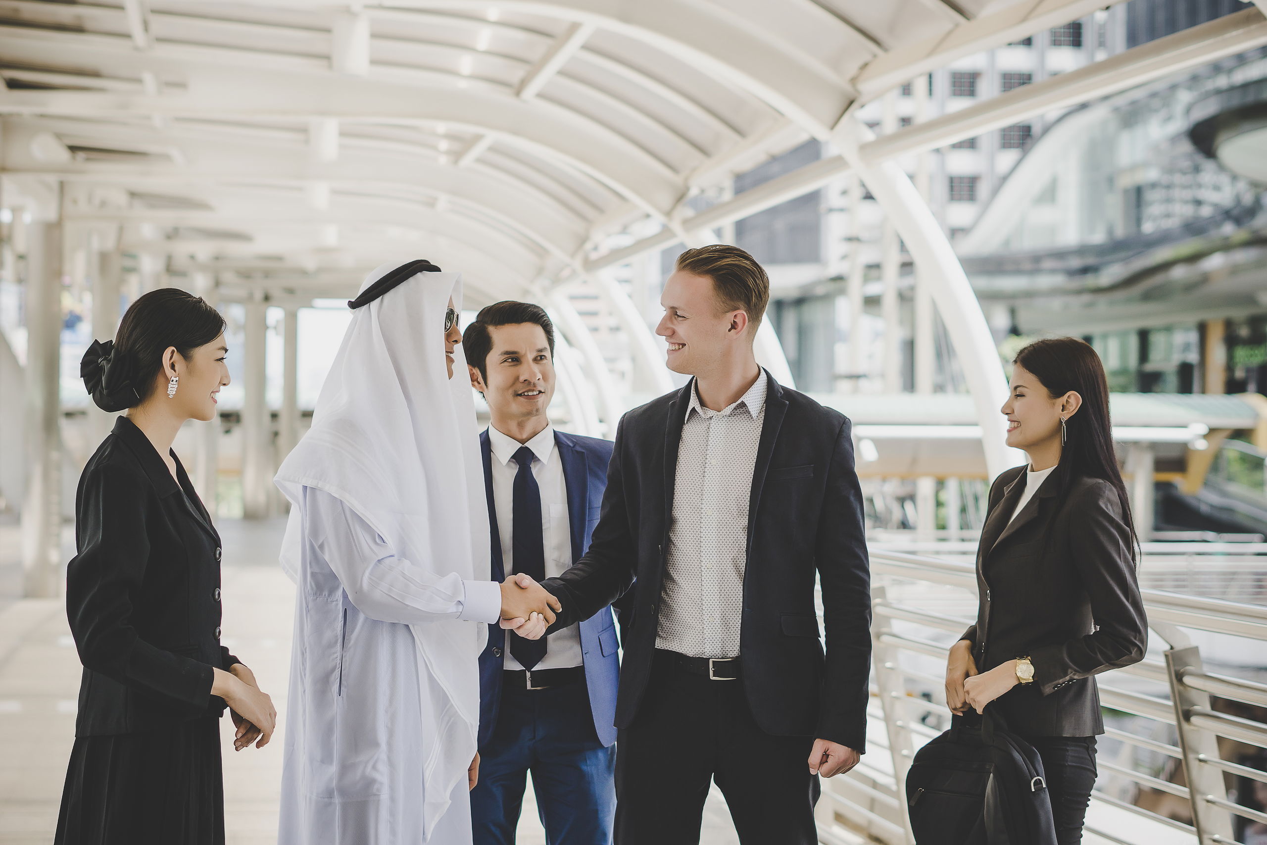 A business delegation meeting on a walkway, a man in Saudi dress shaking hands with a colleague while three others look on